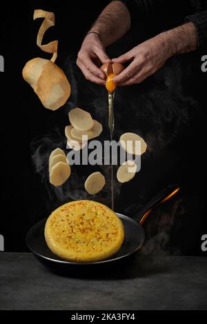 Crop cook breaking egg while half peeled potato with raw pieces dropping on omelette in frying pan Stock Photo