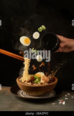 Cook holding noodles with chopsticks and adding miso broth into ramen while eggs and onion with mushrooms falling into bowl Stock Photo