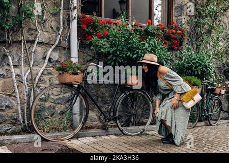 Brunette in romantic dress with bag and hat bending forward and inspecting bike with flowers outside stone house on town street Stock Photo