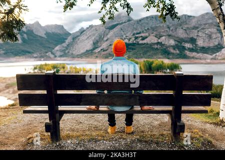 Back view of traveler sitting alone on wooden bench and observing picturesque view of rocky mountains and lake Stock Photo