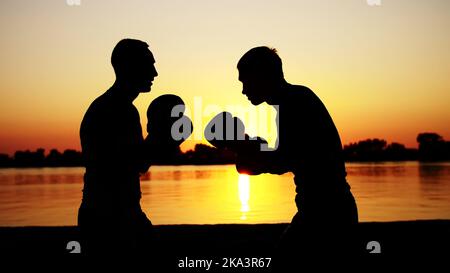 Two dark male figures, at sunrise, against the light, boxing, fighting in sparring, training in a pair of techniques of strikes. On sandy beach, in cargo port, near water, in summer. High quality photo Stock Photo