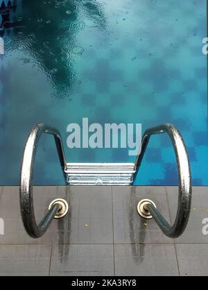 A vertical high angle shot of a metal staircase inside a blue pool with raindrops falling on it Stock Photo