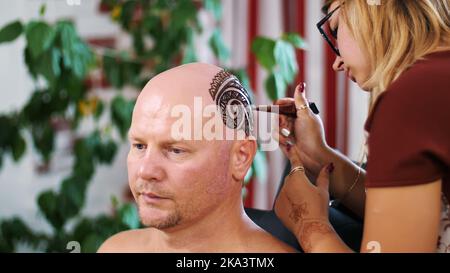 A girl, tattoo master, mehendi artist makes drawing of henna tattoo on scalp of bald Caucasian man, shoulder, neck.The solution of henna dries on the skin. High quality photo Stock Photo