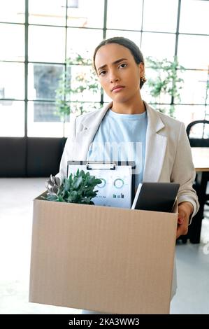 Vertical photo of disappointment upset mixed race woman, stand in her working office, holding a cardboard box with work attributes in her hands, sadly looks at camera, lost her job, fired Stock Photo