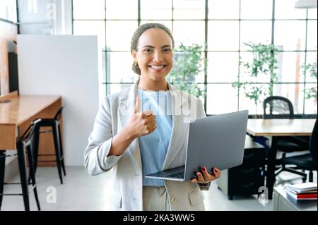 Beautiful positive confident hispanic or brazilian woman, seo, manager, stands in the office in elegant clothes, holds an open laptop in her hand, shows a thumbs-up gesture, looks at camera, smiles Stock Photo