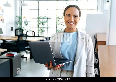 Gorgeous positive confident mixed race successful woman, seo, manager, stands in the office in elegant clothes, holds an open laptop in her hand, looks at camera, smiles friendly Stock Photo