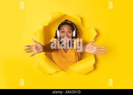 Female Wearing Headphones Stretching Arms Through Hole On Yellow Background Stock Photo