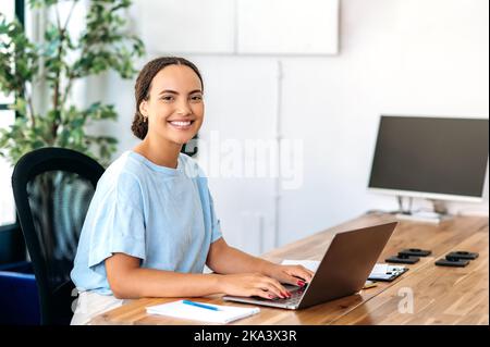 Photo of a successful confident gorgeous mixed race woman, business mentor, financial advisor, programmer, seo, sitting at a work desk in a modern office, looking at camera, smiling friendly Stock Photo
