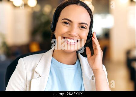 Photo of a positive confident mixed race woman, with headset, call center agent, support service operator, sits in the office, consult clients online, looking at camera, smiling friendly Stock Photo