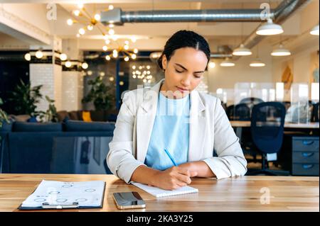 Focused woman takes notes. Mixed race successful woman, business mentor, financial advisor, student, writing information in notebook during online consultation or webinar by video conference Stock Photo