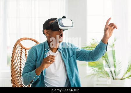 Surprised amazed millennial african american guy in vr glasses plays in virtual game, sits on chair Stock Photo