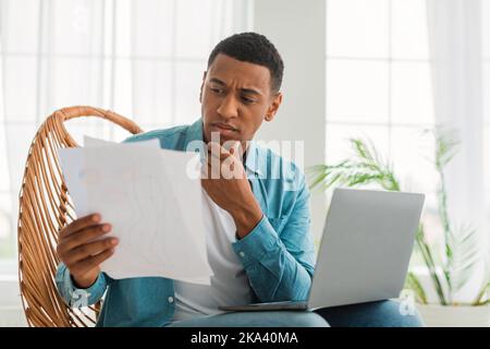 Pensive young african american male working with contract, documents and computer in light living Stock Photo