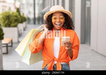 Glad excited shocked young black woman in casual with glasses and hat with open mouth hold many bags, phone Stock Photo