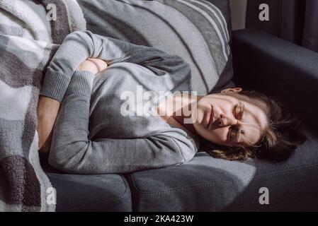 Unhealthy woman struggle with painful ache lying on couch at home. Sad unhappy young female suffering from menstrual pain, feeling sick to stomach, ho Stock Photo