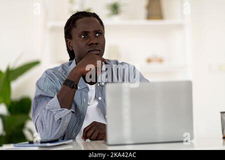 Pensive Young Black Man Sitting On Desk With Laptop And Looking Away Stock Photo