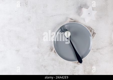 Christmas table setting. Top view of empty grey plate with black cutlery and craft wooden decorations on light table. Event table decoration. Stock Photo