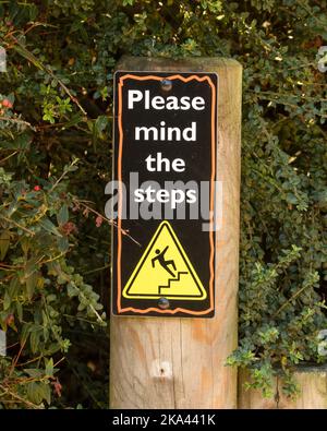 A yellow warning triangle depicting a person slipping with the text 'Please mind the steps' on a black background placed on a wooden pole Stock Photo