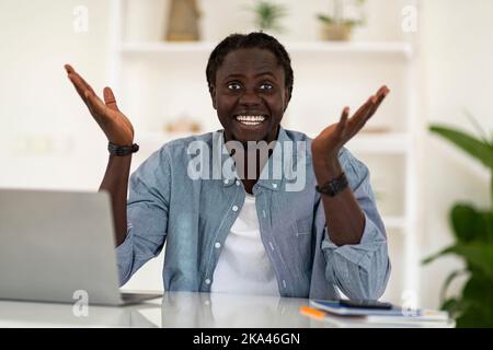 Happy Excited Black Man Sitting At Desk And Spreading Hands At Camera Stock Photo