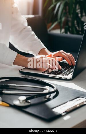 Female doctor in white coat with stethoscope using laptop, writing in medical journal, professional therapist practitioner sitting at table in hospita Stock Photo