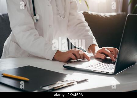 Close up of young woman doctor in white uniform with stethoscope using laptop, writing in medical journal, professional therapist practitioner sitting Stock Photo