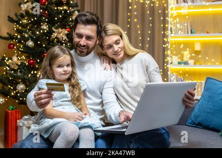 Happy Christmas family doing online shopping sitting at home on sofa, couple husband wife and daughter with laptop and bank credit card having fun choosing Christmas gifts near Christmas tree. Stock Photo
