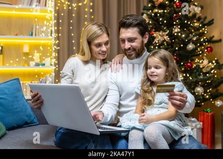 Happy Christmas family doing online shopping sitting at home on sofa, couple husband wife and daughter with laptop and bank credit card having fun choosing Christmas gifts near Christmas tree. Stock Photo
