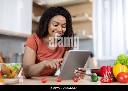 Cheerful young black woman in red t-shirt typing on tablet, read message at table with organic vegetables Stock Photo