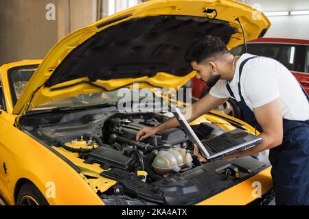 Mechanic man mechanic manager worker using a laptop computer checking car in workshop at auto car repair service center. Engineer young man looking at inspection vehicle details under car hood Stock Photo