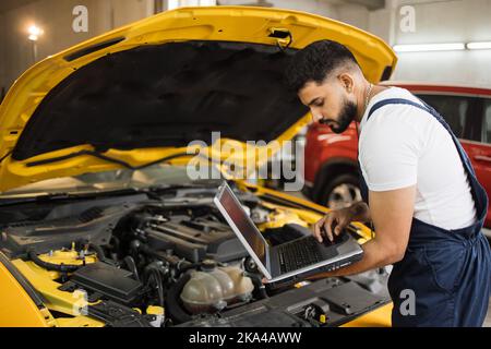 Mechanic man mechanic manager worker using a laptop computer checking car in workshop at auto car repair service center. Engineer young man looking at inspection vehicle details under car hood Stock Photo