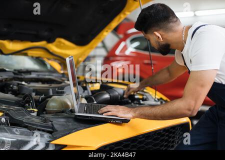 Close up of young bearded engineer mechanic under car hood checking engine with laptop using electronic diagnostic equipment to tune sport yellow vehicle. Stock Photo
