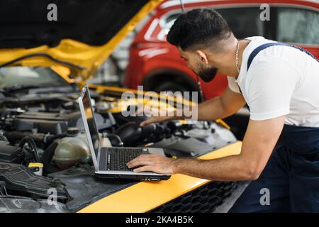 Close up of young bearded engineer mechanic under car hood checking engine with laptop using electronic diagnostic equipment to tune sport yellow vehicle. Stock Photo