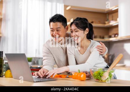 Loving asian spouses using laptop while cooking at kitchen Stock Photo