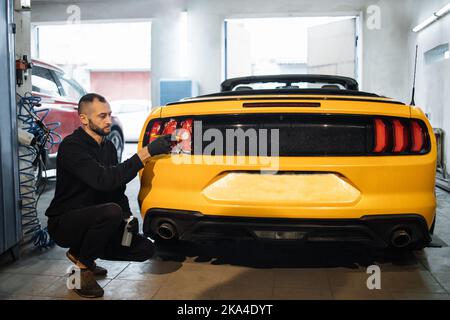 Detailing manual cleaning with soap at car wash. Young male worker in rubber gloves washes car tail light of luxury yellow car with special soap. Car detailing wash Stock Photo