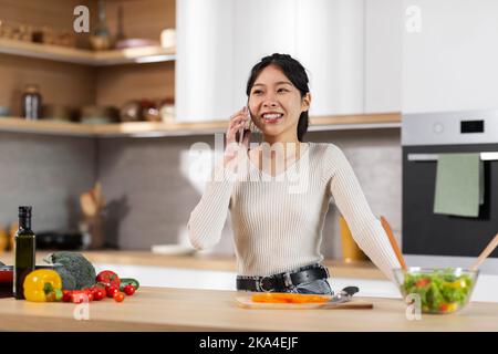 Cheerful pretty asian woman cooking at home, having phone conversation Stock Photo