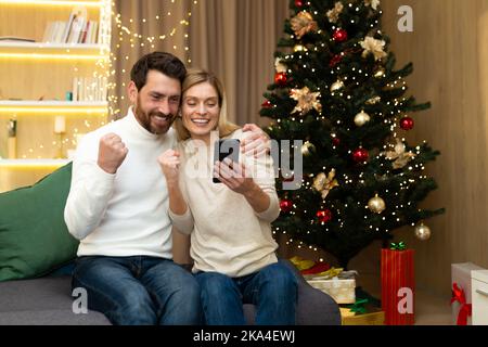 Happy christmas couple man and woman using smartphone smiling and happy celebrating victory discount and shopping in online store near christmas tree Stock Photo