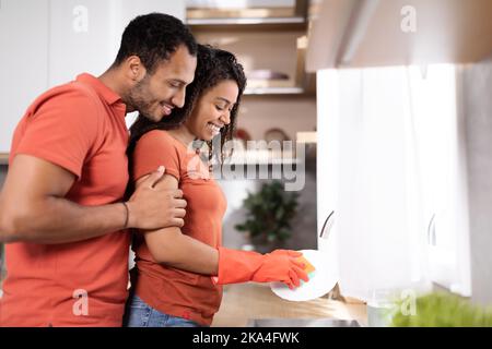 Cheerful young black male hugs wife in rubber gloves, lady washes dishes in kitchen interior, profile Stock Photo