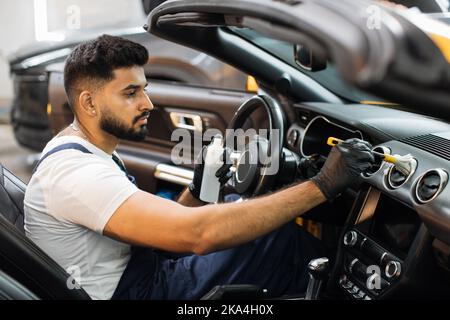Close up portrait of professional car wash service worker in overalls, white t-shirt and protective gloves cleaning car interior and control panel with special chemical anti dust liquid and soft brush Stock Photo