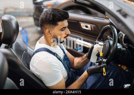 Portrait of young bearded male car detailing worker in overalls and white t-shirt, sitting inside modern vehicle, pouring plastic trim care milk on the soft brush. Car detailing, plastic care. Stock Photo