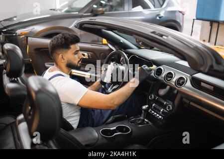 Close up portrait of professional car wash service worker in overalls, white t-shirt and protective gloves cleaning car interior and control panel with special chemical anti dust liquid and soft brush Stock Photo