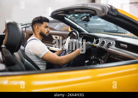 Close up portrait of professional car wash service worker in overalls, white t-shirt and protective gloves cleaning car interior and control panel with special chemical anti dust liquid and soft brush Stock Photo