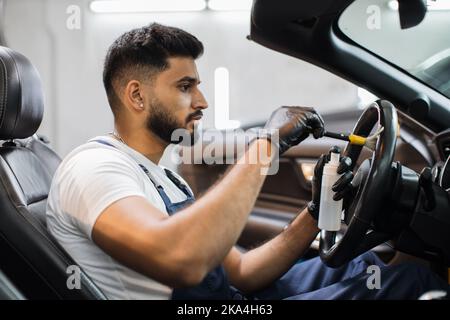 Car detailing and interior care concept. Young man, car wash worker, wearing protective rubber gloves, cleaning the steering wheel of the modern vehicle, using special brush. Stock Photo
