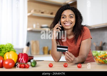 Happy young black female in red t-shirt calling by phone, showing credit card at table with organic vegetables Stock Photo