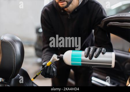 Car detailing and car wash concept. Close up hands of man, car wash worker, wearing black clothes and protective rubber gloves, cleaning car front seat, spraying the foam on the soft brush. Stock Photo