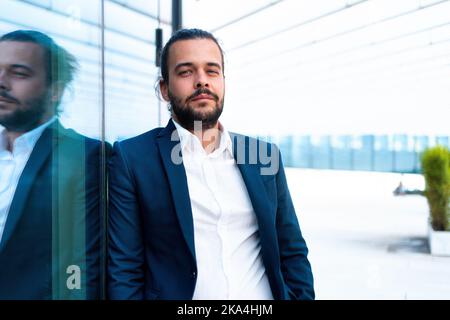 Successful businessman in suit with beard standing near office building confidently looking at camera lean wall. Hispanic male business person portrait. Free space. Modern Business people Stock Photo