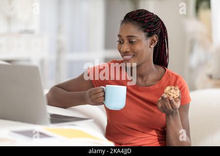 Cheerful Black Woman Relaxing At Home With Laptop And Snacks Stock Photo