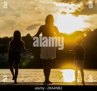 The silhouettes of the mother with children standing on the shore and enjoying the beautiful sunset. Stock Photo