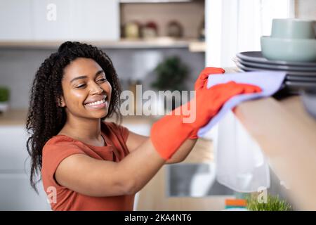 Cheerful pretty young black woman in red t-shirt and rubber gloves dusts off from wooden shelf Stock Photo