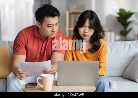 Upset asian couple checking bills, using laptop, home interior Stock Photo