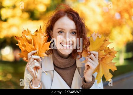 Smiling pretty young european red-haired lady in raincoat hold yellow leaves, has fun alone in city park Stock Photo