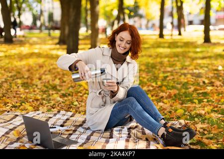 Cheerful pretty young european red-haired female in raincoat with computer pours drink from thermos Stock Photo